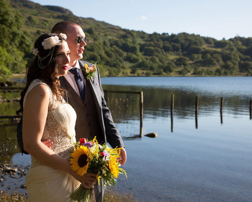 Couple portrait at golden hour beside a calm lake, framed by trees with reflections in the water and a sunflower bouquet glowing in the evening sun. Captured by Tim Christian Jones | Leeds wedding photographer.