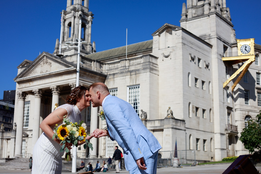Couple portrait of Gemma and Danny sharing a playful moment beside piano festival art on Millennium Square in Leeds, with Leeds Civic Hall in the background on a bright blue sky wedding day
